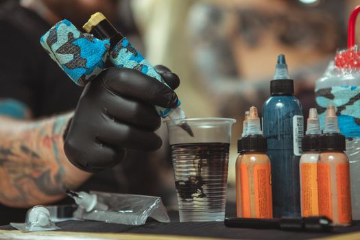 Cropped close up of tattoo ink spreading in glass of water, tattoo artist cleaning his tattoo machine in a glass of water, while working. Selective focus on tattoo machine of a tattooist, copy space