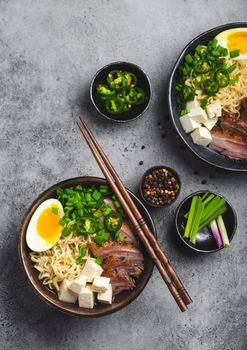 Two bowls of tasty Asian noodle soup ramen with meat broth, tofu, pork, egg with yolk on grey rustic concrete background, close up, top view. Hot tasty Japanese ramen soup for dinner asian style