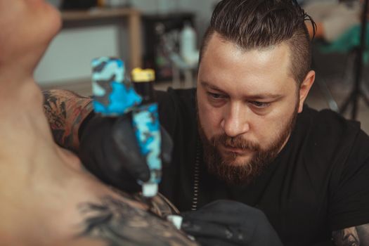 Professional tattoo artist looking focused, working at his studio, making a tattoo on the chest of his client. Bearded tattooist making a tattoo for his client