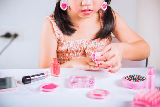 Asian adorable funny little girl making makeup dips brush into bottle to paints nails polish red nail varnish herself, Learning activity to be woman, happy kid is beautiful make up with cosmetics toy