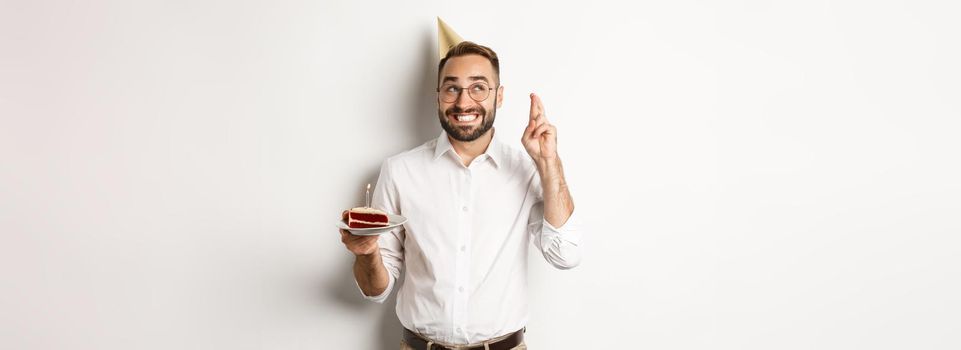 Holidays and celebration. Happy man having birthday party, making wish on b-day cake and cross fingers for good luck, standing against white background.