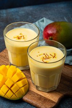 Healthy Indian Ayurveda drink mango lassi in two glasses on rustic concrete table with fresh ripe cut mango, yellow blended beverage made of mango fruit, yoghurt or milk curd and spices