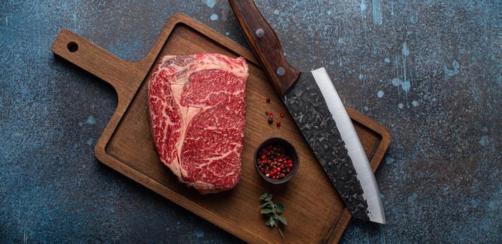 Raw meat beef marbled prime cut steak Ribeye on wooden cutting board and rustic concrete kitchen table background from above with big knife and spices, beefsteak concept