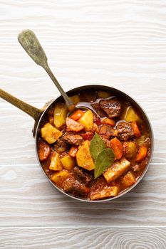 Delicious stew with meat, potatoes, carrot, herbs and gravy in rustic copper pot on white wooden background from above. Traditional winter and autumn dish beef and vegetables ragout in stewpot