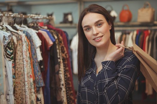 Beautiful dark haired young woman smiling to the camera, holding shopping bag, standing at clothing store. Charming female customer carrying her purchase at fashion shop, copy space