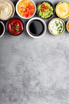 Set of different healthy sauces in bowls on rustic concrete background, top view, close-up. Space for text. Tomato ketchup, mayonnaise, guacamole, mustard, pesto, cheese sauce - assortment of dips