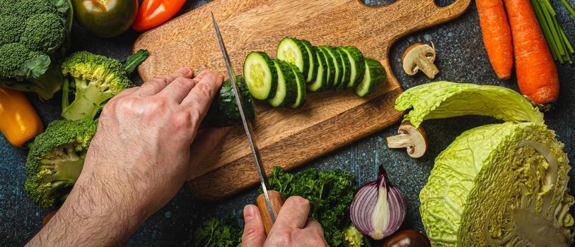 White man hands chopping cucumber on wooden cutting board with knife and assorted fresh vegetables on rustic concrete table, diet and vegetarian food preparing, meal cooking concept, healthy lifestyle