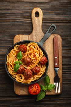 Top view of delicious pasta with meatballs, tomato sauce and fresh basil in cast iron rustic vintage pan served on cutting board, wooden background. Tasty homemade meatballs spaghetti