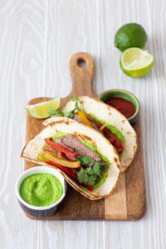 Traditional Mexican dish Beef fajitas tacos served on wooden cutting board with tomato salsa and guacamole on rustic white wooden background from angle view, American Mexican food