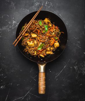 Stir fry soba noodles with chicken, vegetables in old rustic wok pan, chopsticks on black stone background, close up, top view. Traditional asian/thai meal, close up