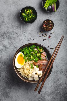 Delicious Asian noodle soup ramen in bowl with meat broth, tofu, sliced pork, egg with yolk, grey rustic concrete background, close up, top view. Hot tasty Japanese ramen soup for dinner asian style