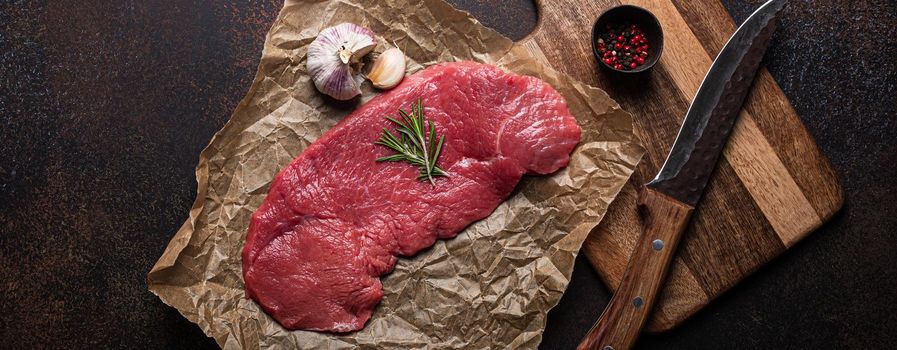 Beef lean raw fillet steak on baking paper with rosemary, garlic and spices on dark brown rustic concrete background with knife from above flat lay, diet beef meat steak ready to be cooked