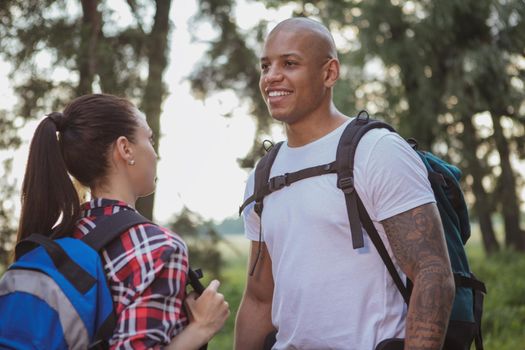 Handsome African man laughing, enjoying travelling with his girlfriend. Couple backpacking in the forest. Attractive male traveler talking to his woman while resting during hiking