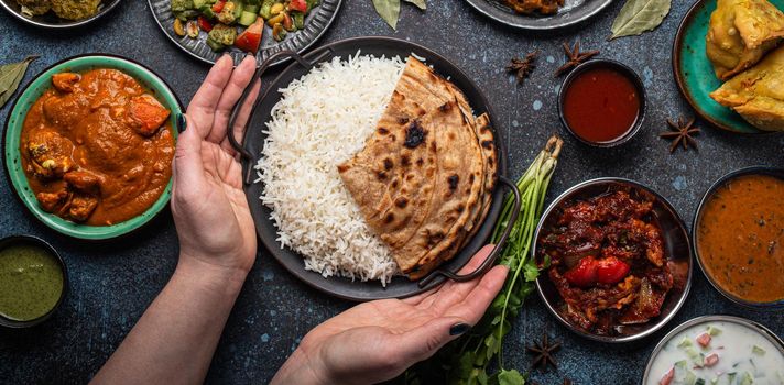Female hands serving Indian ethnic food buffet on rustic concrete table from above: curry, fried samosa, rice biryani, dal, paneer, chapatti, naan, chicken tikka masala, dishes of India for dinner
