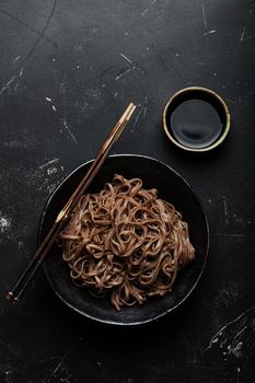 Bowl with plain soba noodles on dark black stone background from above, noodles for cooking Chinese Thai or Japanese dish with soy sauce