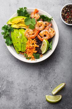 Close-up, top view of salad bowl with shrimps, avocado, fresh kale, quinoa, red lentils, lime and olive oil on gray stone background. Lunch bowl, healthy clean eating, dieting or nutrition concept