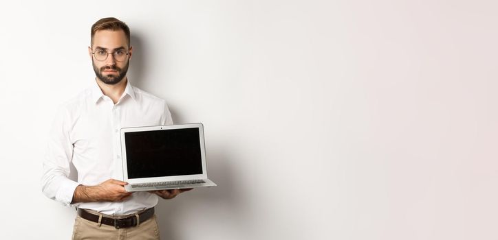 Confident manager demonstrating presentation on screen, showing laptop display and looking serious, standing over white background.