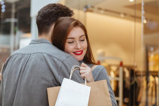 Adorable young loving couple hugging at the shopping mall, copy space. Gorgeous woman smiling with her eyes closed, embracing her boyfriend, holding shopping bags. Presents, anniversary, valentines concept