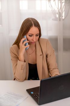 European professional woman is sitting with a laptop at a table in a home office, a positive woman is studying while working on a PC. She is wearing a beige jacket and jeans and is on the phone