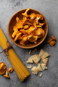 Wild forest mushrooms chanterelles in wooden bowl, raw pasta spaghetti, cheese parmesan on concrete rustic background top view. Seasonal autumn food, ingredients for cooking fresh chanterelles pasta