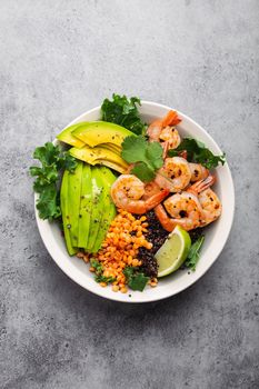 Close-up, top view of salad bowl with shrimps, avocado, fresh kale, quinoa, red lentils, lime and olive oil on gray stone background. Lunch bowl, healthy clean eating, dieting or nutrition concept