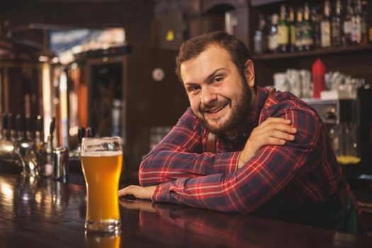 Friendly bearded bartender smiling joyfully to the camera, enjoying working at his beer pub, copy space. Happy brewer serving delicious beer, leaning on a counter. Service, lifestyle concept