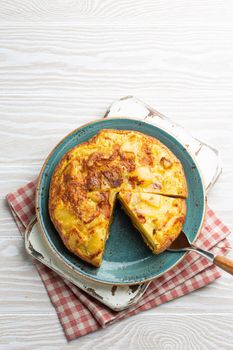 Homemade Spanish tortilla with one slice cut - omelette with potatoes on plate on white wooden rustic background top view. Traditional dish of Spain Tortilla de patatas for lunch or snack, overhead.