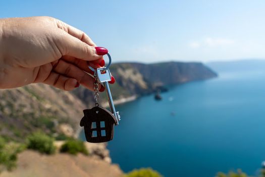 Keys with a keychain in the shape of a house in a female hand against the backdrop of the sea. The concept of buying a house, apartment, real estate, young family, mortgage loan, rental housing