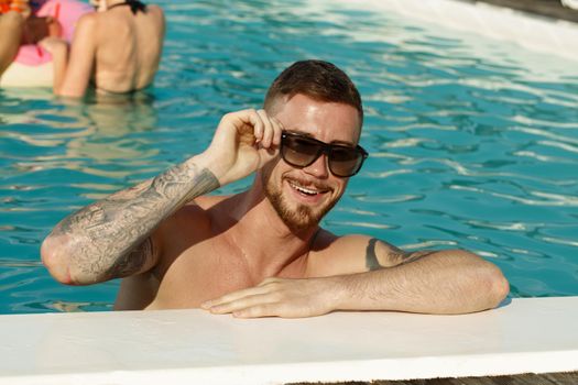 Handsome cheerful bearded man wearing sunglasses smiling to hte camera while relaxing at the swimming pool, copy space. Happy man enjoying hot summer day at the pool. Vacation, lifestyle concept