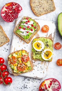 Avocado toasts with egg, tomatoes, seasonings on white wooden rustic cutting board, stone background. Ingredients for healthy breakfast avocado sandwiches with different toppings, top view, close-up.