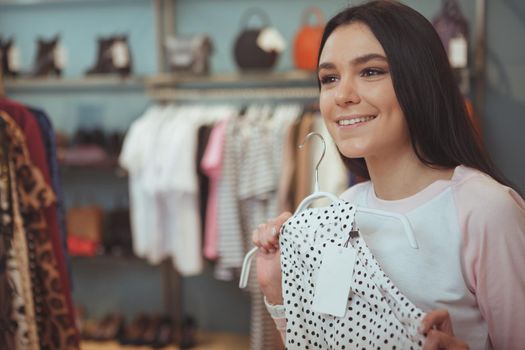 Beautiful happy dark haired woman smiling, trying a new dress at fashion boutique. CHeerful young female customer choosing new dress to buy during seasonal sales, copy space