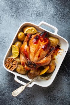 Close-up of baked or roasted whole juicy crispy organic chicken with potatoes, garlic, lemon in white casserole dish with serving spoon on concrete rustic background, top view