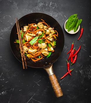 Stir fry chicken with vegetables in old rustic wok pan, chopsticks on black stone background, close up, top view. Traditional asian/thai meal
