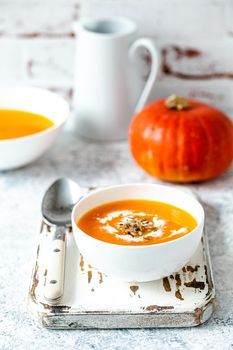 Homemade pumpkin cream soup served in white ceramic bowl on white table with spoon decorated with whole pumpkin, angle view, selective focus. Autumn cozy comfort food .