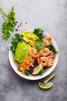 Close-up, top view of salad bowl with shrimps, avocado, fresh kale, quinoa, red lentils, lime and olive oil on gray stone background. Lunch bowl, healthy clean eating, dieting or nutrition concept