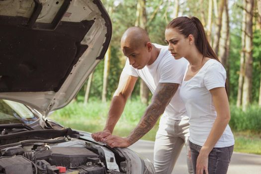 Young multiracial couple examining their broken down car on a countryside road. African handsome man and his girlfriend waiting for roadside assistance near their broken car, during roadtrip