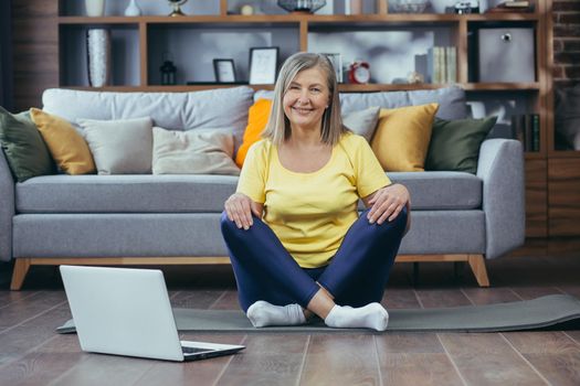 Portrait of active senior woman sitting at home on lotus floor and looking at camera, happy grandmother doing yoga online using laptop