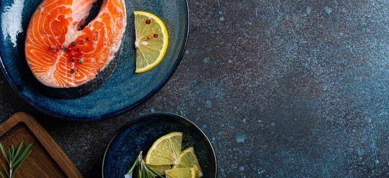 Uncooked raw fresh fish salmon steak top view on plate rustic dark concrete stone background with rosemary, lemon wedges and spices, delicacy healthy fish cooking and nutrition concept space for text