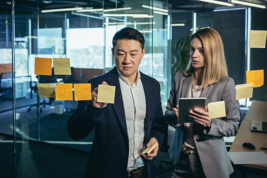 Two employees, writing a plan and tasks on the task board, an Asian man and a woman, working in a modern office, write tasks on colored notes