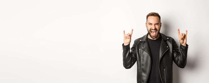 Cool adult man in black leather jacket, showing rock on gesture and tongue, enjoying music festival, standing over white background.