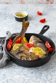 Roasted or grilled fish sea bream or dorado with lemon, herbs and tomatoes in cast iron skillet great for healthy meal or Mediterranean diet on rustic white stone table, angle view