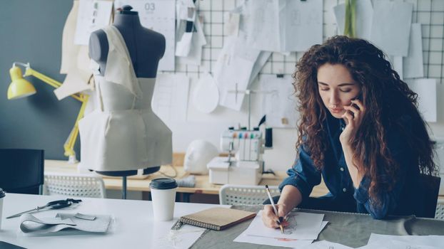 Professional female fashion designer is talking on mobile phone and drawing clothing sketch with pencil at table in tailoring shop. Everyday work and communication in workplace concept.