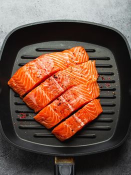 Top view, close-up of cut in slices fresh raw salmon fillet with seasonings on grill skillet, gray stone background. Preparing salmon fillet for cooking, healthy eating concept