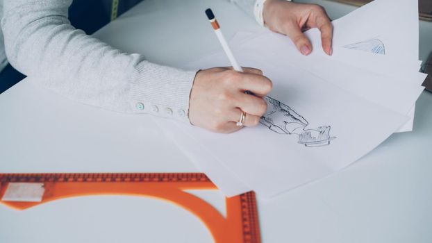 Close-up shot of young woman's hand drawing outlines of stylish ladies' garment with attention. Paper, tailor's scissors, ruler are visible on studio table. Designer at work concept.