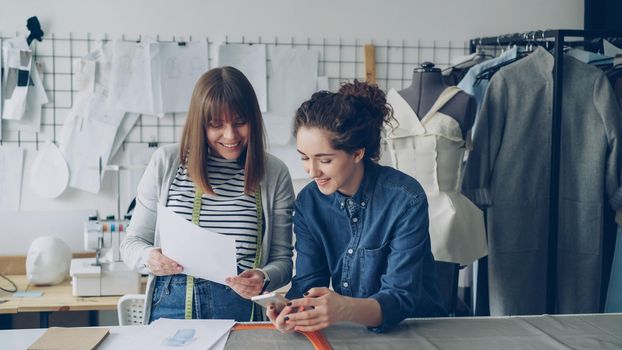 Creative dressmakers are using smartphone and looking at clothing sketches while working in nice modern studio. Dummy, sewing machine and women's garments in background.