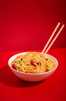 Asian Chinese noodles with shrimps and vegetables in white bowl with wooden sticks on red minimal paper background, hard sharp shadows, minimal foodphoto concept