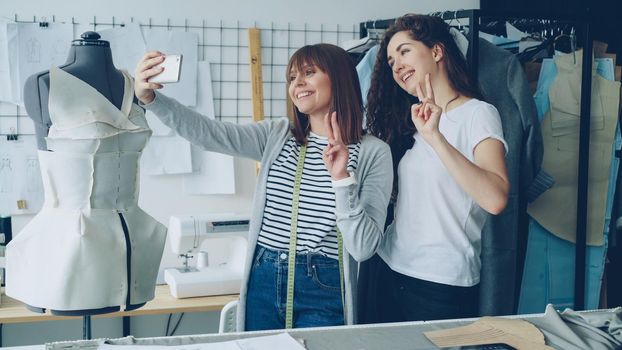 Two cheerful female clothing designers are making funny selfie with smart phone while standing beside clothed mannequin in studio. Attractive women are smiling and posing.