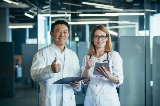 Team of two doctors asian man and woman looking at camera and smiling holding thumbs up working in modern clinic