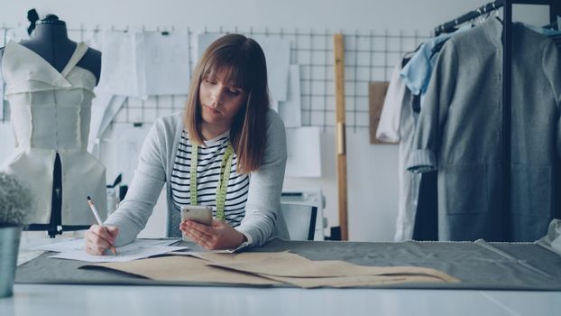 Creative clothing designer is looking at smartphone screen and drawing sketch while working in modern tailor shop at sewing table. Woman is busy and involved in process.