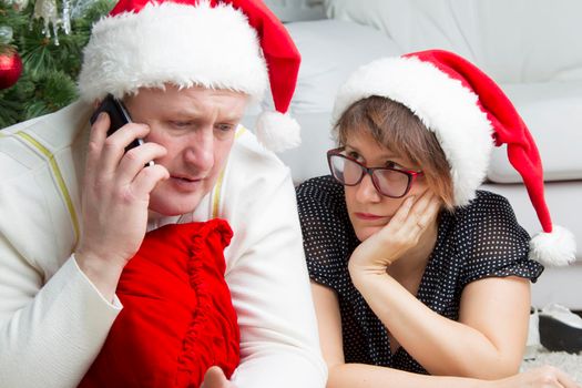 Husband and wife on New Year's Eve. Problems in Christmas. A man with a cell phone and a worried woman in funny Santa Claus hats.A married couple on holidays is preoccupied